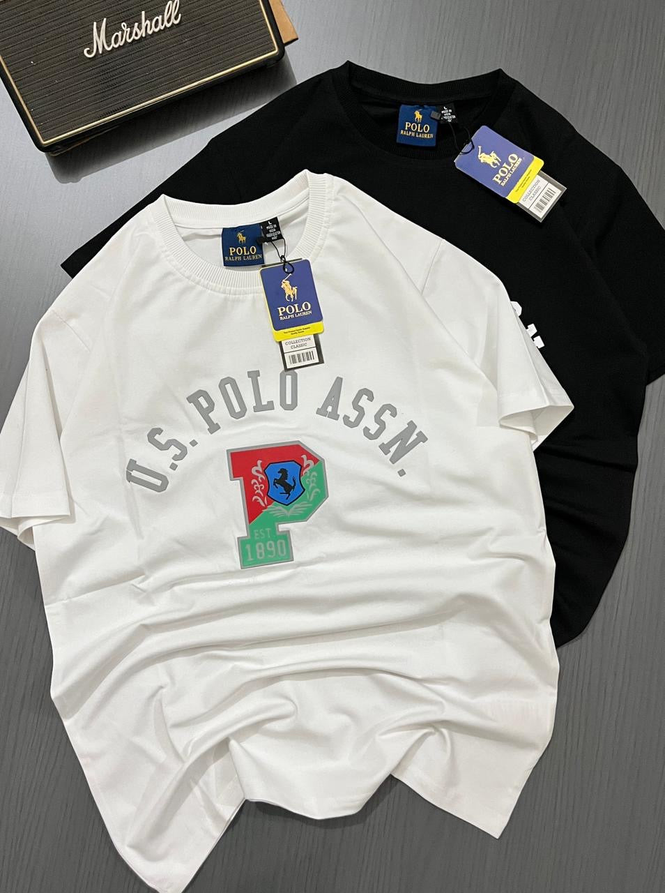 White and black t-shirts with U.S. Polo Assn. branding on a gray surface.